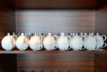 Fototapeta na wymiar Many white stacked porcelain tea cups and small teapots in a dark brown wooden shelf. Teapot and cup, set for one person, consisting of a small white teapot and a teacup.