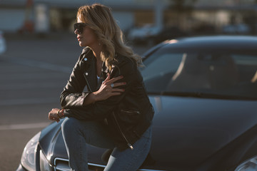 Fototapeta na wymiar Photo of young blonde woman in sunglasses sitting on hood of black car
