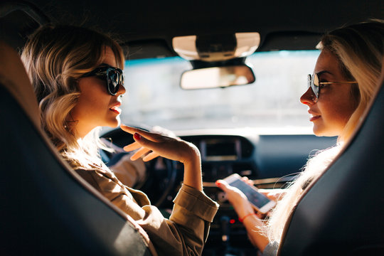 Photo of back of two blonde women with long hair and phone in hand sitting in car