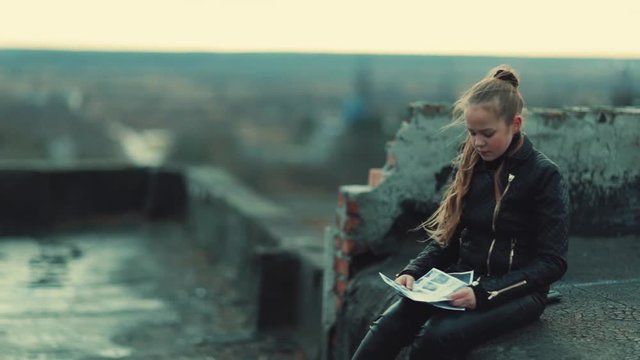 Upset little girl with long ponytail sitting on roof, holding papers in hands, reading. Brooding schoolgirl. Unrequited love. Outdoors. Depression. Bad mood.