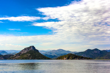 The beautiful smooth surface of the blue lake reflects the blue sky with white clouds, hilly terrain with trees among the stone slopes on the shore. Skadar Lake, Podgorica region, Montenegro.