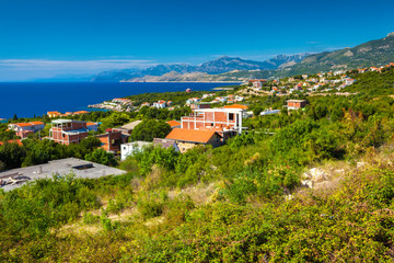 Fototapeta na wymiar Brick houses on the beautiful green coast of the blue Adriatic Sea against the backdrop of magnificent mountains on the horizon on a sunny summer day. View from the highway, Ulcinj, Montenegro.