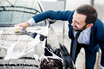 Elegant businessman dressed in the suit washing his car with yellow sponge and foam on a self service car wash