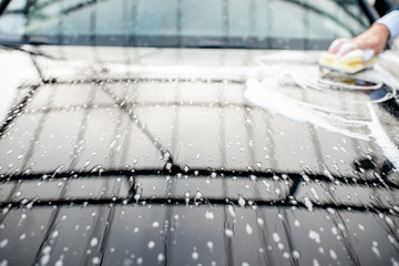 Close-up of a car hood during the washing process with yellow sponge and foam outdoors