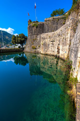 Large historical medieval stone wall in front of a moat with green water on the backdrop of the picturesque mountains on a sunny day. Kotor Venetian fortifications, Kampana Tower Old Town, Montenegro.