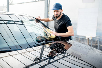 Professional washer in t-shirt and cap wiping windshield with yellow microfiber at the open air car...