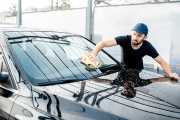 Professional washer in t-shirt and cap wiping windshield with yellow microfiber at the open air car wash