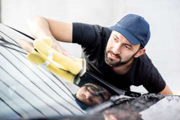 Professional washer in t-shirt and cap wiping windshield with yellow microfiber at the open air car wash