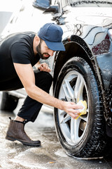 Professional washer in black uniform and cap wiping with sponge car wheel during the washing process outdoors