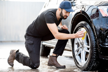 Professional washer in black uniform and cap wiping with sponge car wheel during the washing...