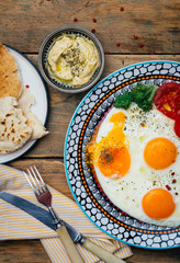 Breakfast table setting. Fried eggs on a plate with tomatoes, bread (pita) and hummus on a wooden table. Top view. Wooden background.