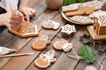 Icing of Christmas bakery. Woman decorating honey gingerbread cookies on wooden brown table. closeup, copy space. Blank biscuit gingerbread house, ready to decorate.