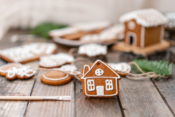 Cute little house in the foreground. A lot of ginger biscuits in different form on brown wooden table. Decorated with white sweet glaze. Christmas mood, winter morning. Fir branches