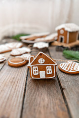 Cute little house in the foreground. A lot of ginger biscuits in different form on brown wooden table. Decorated with white sweet glaze. Christmas mood, winter morning. Fir branches