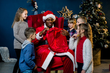 African Santa Claus sits on a background of a Christmas tree and presents gifts to the children