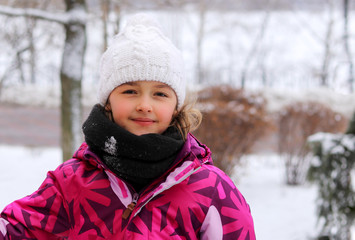 Outdoor portrait of cute little girl in white knitted hat in winter park smiling looking at camera....