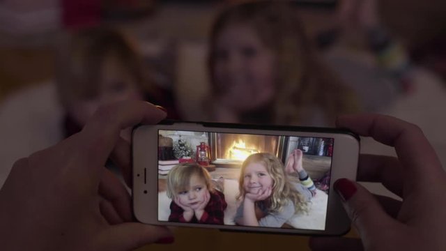 Mom Takes A Candid Video, Using Smart Phone, Of Her Kids By Cozy Fire At Christmas Time, Little Boy Looks Bored, His Sister Looks Happy