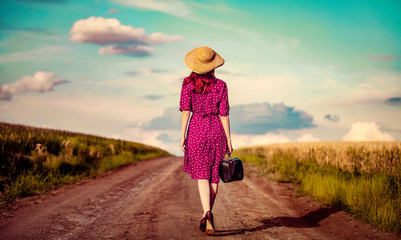 Redhead girl in red dress with hat and suitcase walking down on rural road near wheat field and...