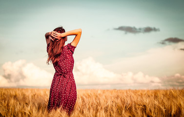 Style redhead girl in red dress tay on yellow wheat field in sunset time and looking forward. Image...