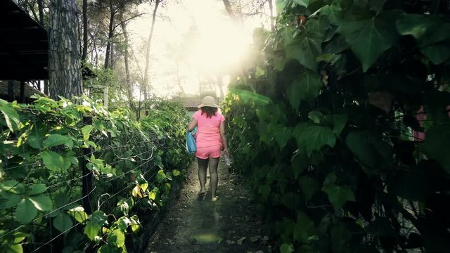 Tracking two woman walking to beach on narrow foliage passage amoung camping wooden houses in the forest, cinematic steadicam shot