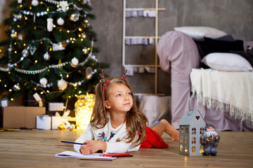 Portrait of Little cute girl writing a letter for Santa near Christmas tree on wooden floor at home. Happy new year.