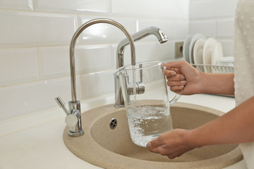 Woman pouring water into glass jug in kitchen, closeup