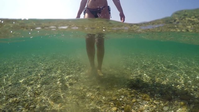 Sexy fashion model in bikini walking in shallow sea water toward camera on beach sith stones, gopro dome shot