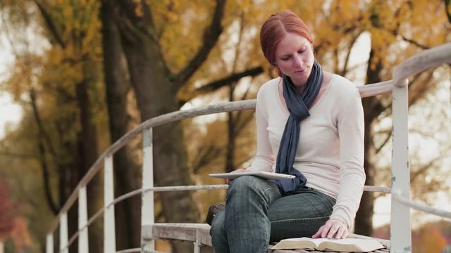 A young Christian woman studying the Bible with a tablet in a park by autumn.