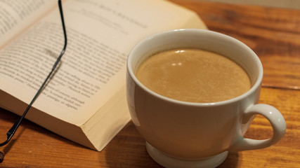 A white ceramic cup of coffee with cream next to a book and reading glasses, sitting atop a wooden coffee table