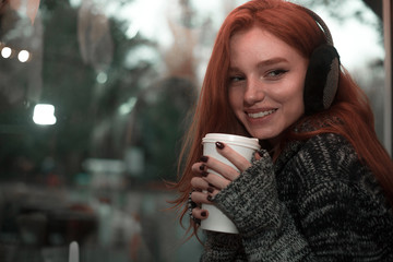 The portrait of a redhead smilling girl with cup of coffee in gray sveater sitting in a cafe pleased