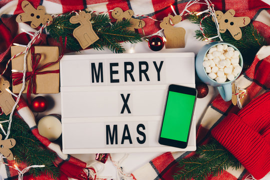 Merry Xmas Text Displayed On A Lightbox With Smartphone With Green Screen Surrounded Christmas Decoration With Fir Branches, Red Plaid And Lights. Flat Lay. Top View. Chroma Key