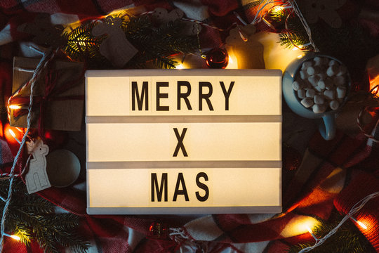 Merry Xmas text displayed on a lightbox with beautiful Christmas decoration with fir branches, red plaid and lights in the dark. Flat lay. Top view