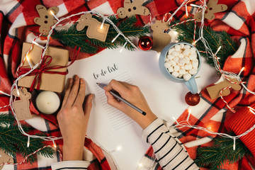Woman hands writing new year to do list on white piece of paper, planning her future. Christmas decoration with red details. Flat lay. Top view. Mockup, copy space for your text.