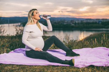 Beautiful pregnant woman enjoys drinking water while spending time outdoor.