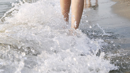 Closeup of female barefoot legs walking on sea coast being splashed by waves, cinematic steadicam...