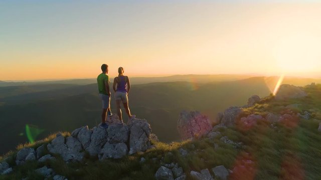 AERIAL, LENS FLARE: Flying near hiker couple watching the sunset after reaching the summit of a grassy hill in the Slovenian countryside. Sporty couple observing the picturesque evening landscape .