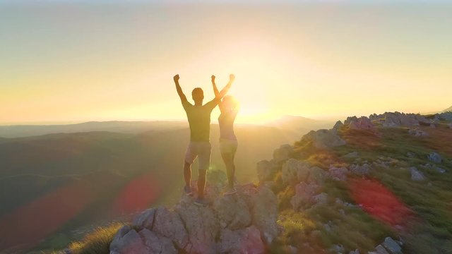 LENS FLARE, DRONE: Excited Caucasian couple celebrates reaching top of the grassy mountain at sunset. Picturesque view of the beautiful summer nature surrounding excited young hikers pumping fists.