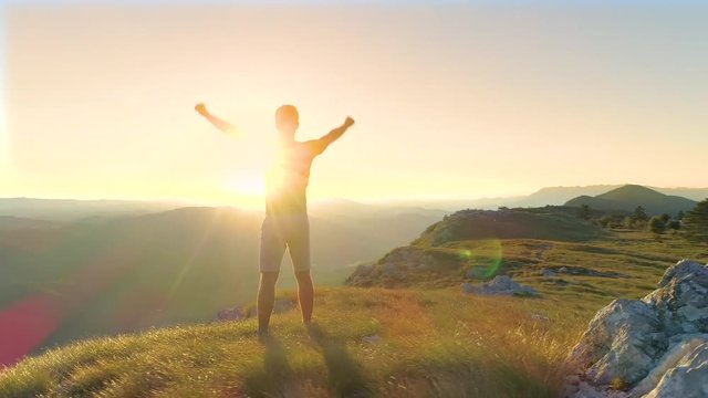 AERIAL, LENS FLARE: Cheerful Caucasian man outstretches arms victoriously while observing the sunset. Happy male hiker observing the picturesque valley on a sunny summer morning. Carefree trekker.