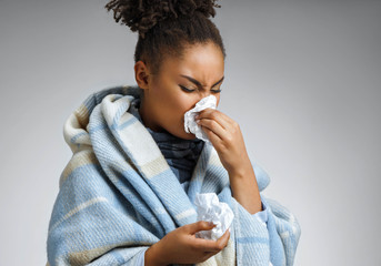 Sick woman sneezing and blowing nose. Photo of african american woman wrapped in paid on gray...