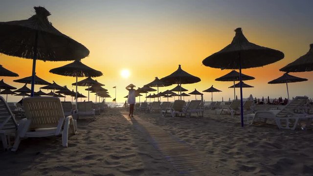 Woman in white dress and hat walk toward camera on beach in the seaside resort with sun loungers and wooden sheds against sunset, cinematic