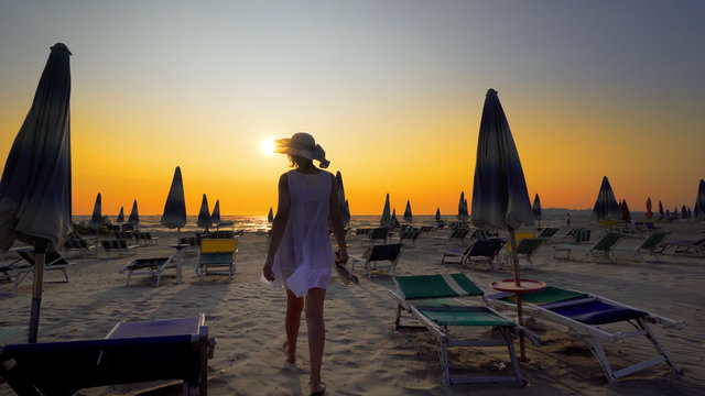 Happy woman in white dress and hat walking and turning on beach at sunset. Seaside resort with sun loungers and wooden sheds against sunset, cinematic