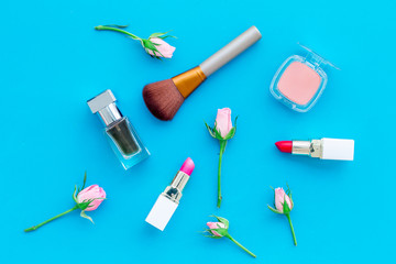 Rose tone cosmetics. Lipstick, bulk, eyeshadow, perfume among rose flowers on blue background top view
