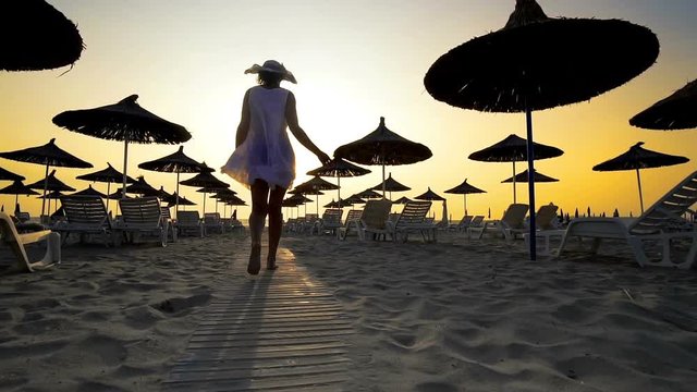 Happy woman in white dress and hat walking and turning on beach at sunset. Seaside resort with sun loungers and wooden sheds against sunset, cinematic slow motion