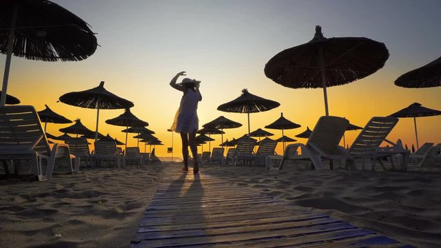 Happy woman in white dress and hat walking and turning on beach at sunset. Seaside resort with sun loungers and wooden sheds against sunset, cinematic