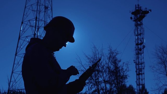 Silhouette of an engineer at sunset, who looks at the telecommunication pillars. Engineer writes data to the tablet. The concept of modern telecommunications