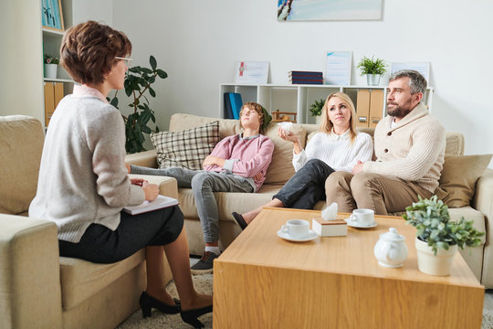 Tired parents sitting on sofa and discussing behavior of teenage son with psychologist sad mother crying and holding paper napkin while sharing her emotions at therapy session.