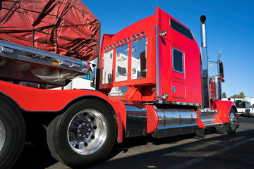 Bright red big rig classic American semi truck with flat bed semi trailer carry commercial cargo covered and fixed by slings stand on truck stop