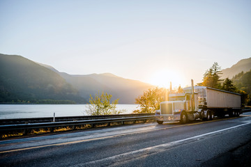 Classic big rig semi truck with bulk semi trailer driving on the road along the river in sunlight