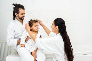 Obraz premium Parents sitting in bathroom with toddler son in white bathrobe