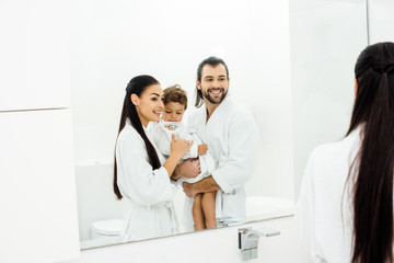 Mom and dad looking in mirror and holding toddler son in white bathrobes